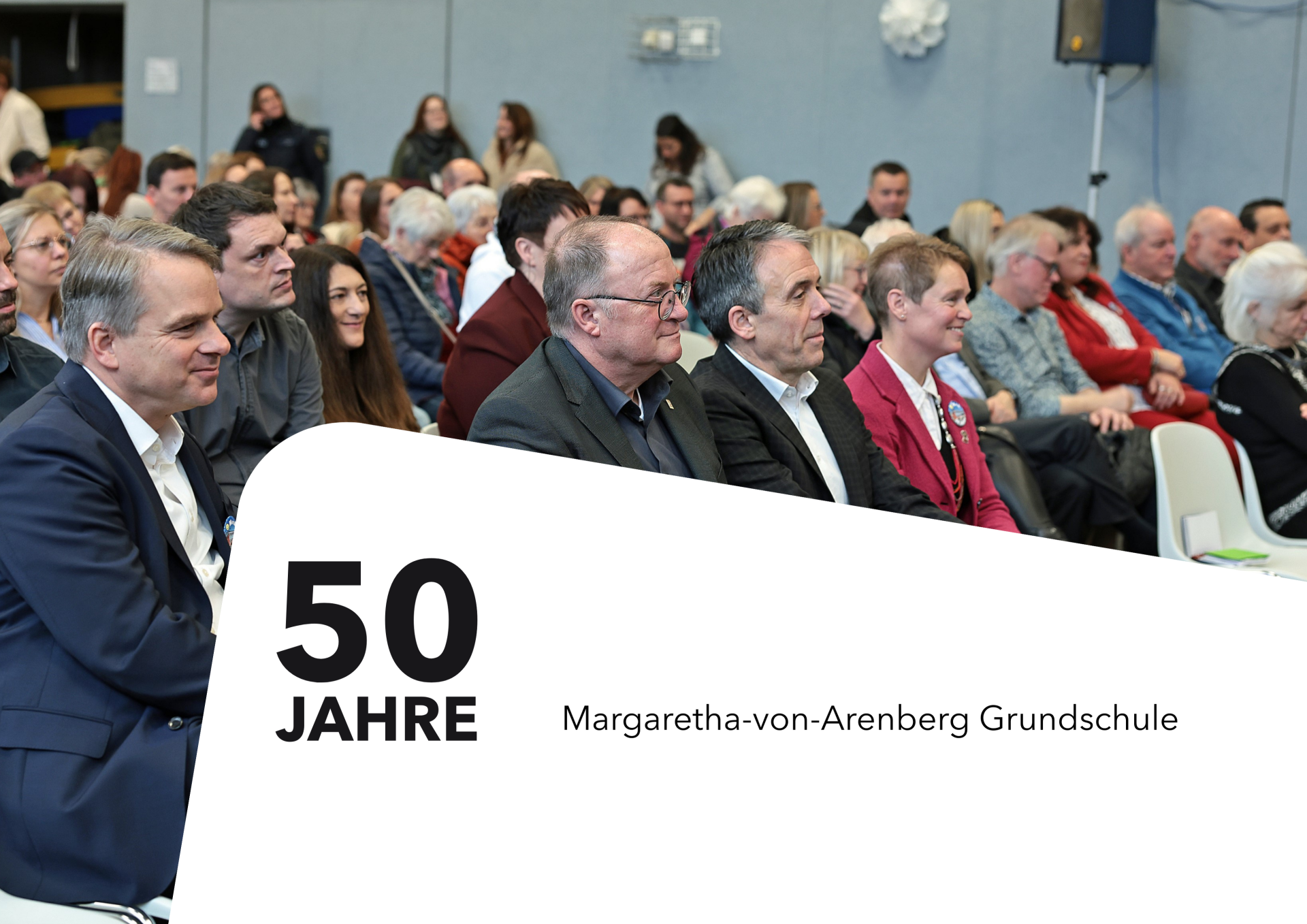 Publikum bei der Feier zum 50-jährigen Bestehen der Margaretha-von-Arenberg-Grundschule sitzt aufmerksam im Veranstaltungsraum. In der ersten Reihe befinden sich Ehrengäste. Im Bild steht der Text „50 Jahre Margaretha-von-Arenberg Grundschule“.