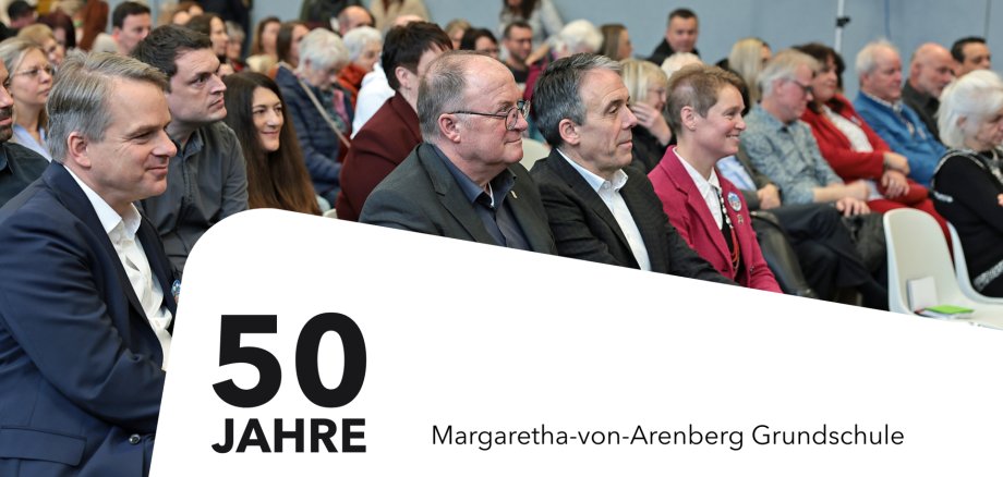 Publikum bei der Feier zum 50-jährigen Bestehen der Margaretha-von-Arenberg-Grundschule sitzt aufmerksam im Veranstaltungsraum. In der ersten Reihe befinden sich Ehrengäste. Im Bild steht der Text „50 Jahre Margaretha-von-Arenberg Grundschule“.