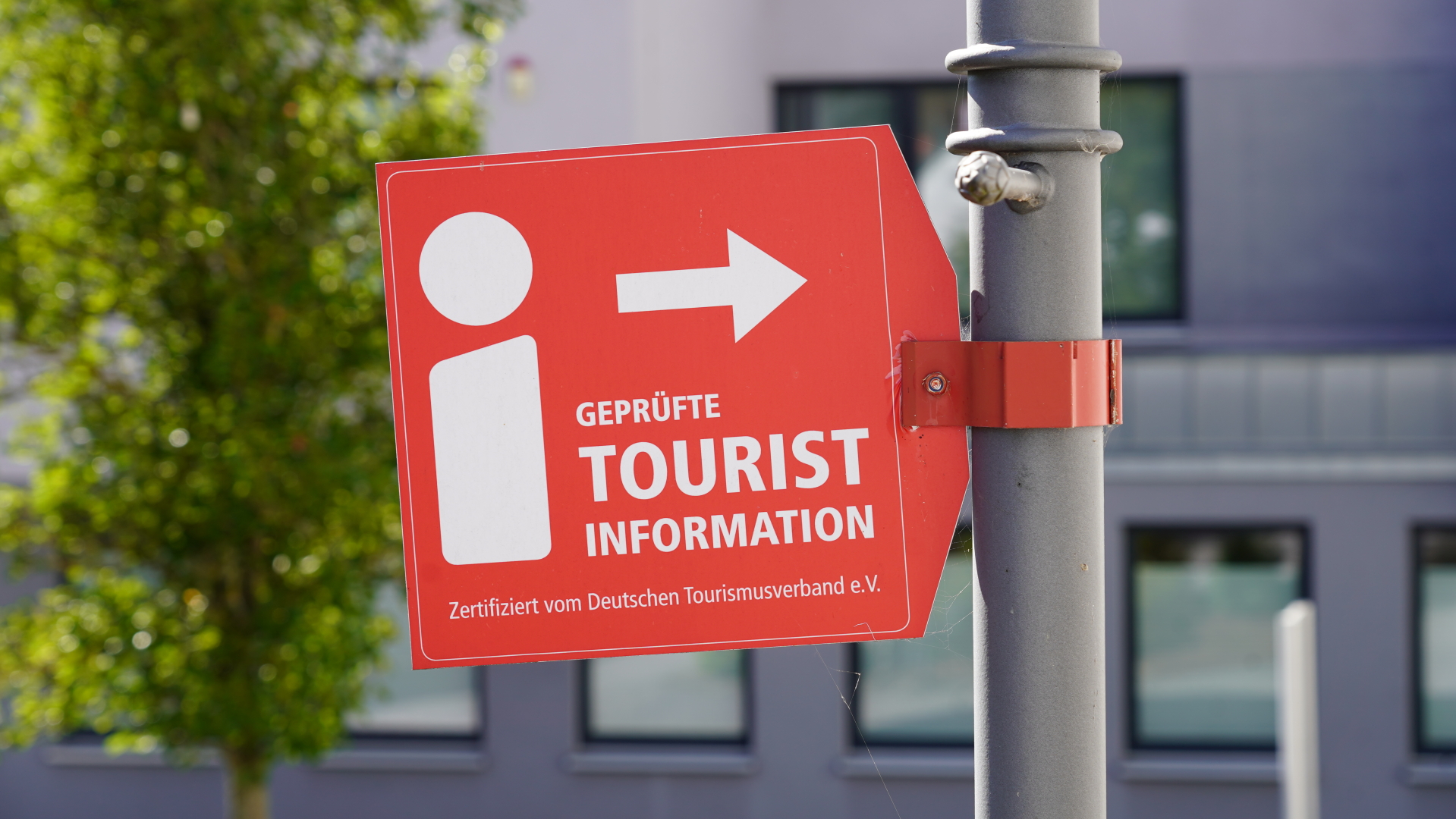 Ein rotes Schild, das an einem grauen Pfosten befestigt ist. Das Schild zeigt ein weißes Informationssymbol und einen Pfeil nach rechts. Darunter steht „Geprüfte Tourist Information“ und „Zertifiziert vom Deutschen Tourismusverband e.V.“