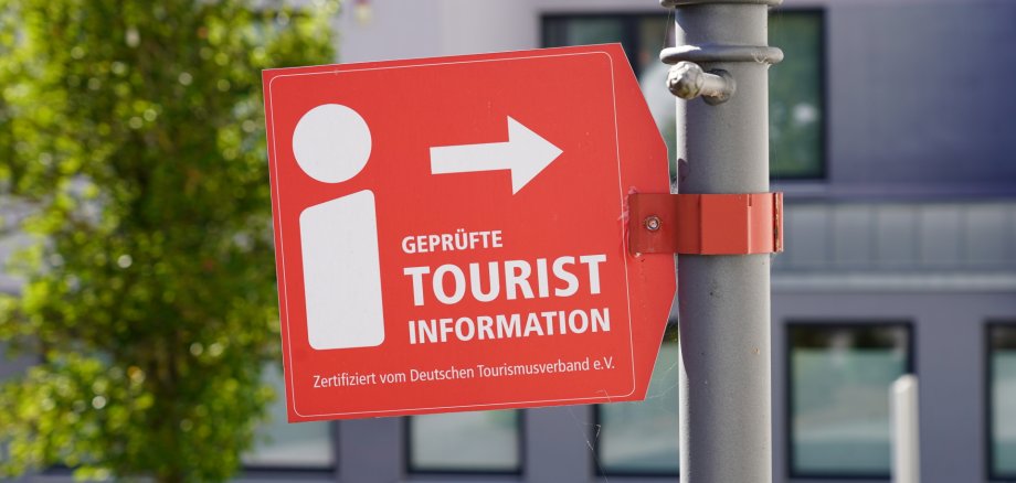 Ein rotes Schild, das an einem grauen Pfosten befestigt ist. Das Schild zeigt ein weißes Informationssymbol und einen Pfeil nach rechts. Darunter steht „Geprüfte Tourist Information“ und „Zertifiziert vom Deutschen Tourismusverband e.V.“