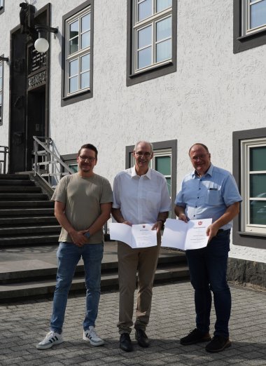 In der Mitte hält Bernhard Jüngling ein Dokument mit einem Wappen, das ihm zu seinem 40-jährigen Dienstjubiläum überreicht wurde. Bürgermeister Guido Nisius steht rechts neben ihm und gratuliert. Links steht Tim Schneider vom Personalrat. Im Hintergrund ist die Verbandsgemeindeverwaltung Adenau zu sehen.