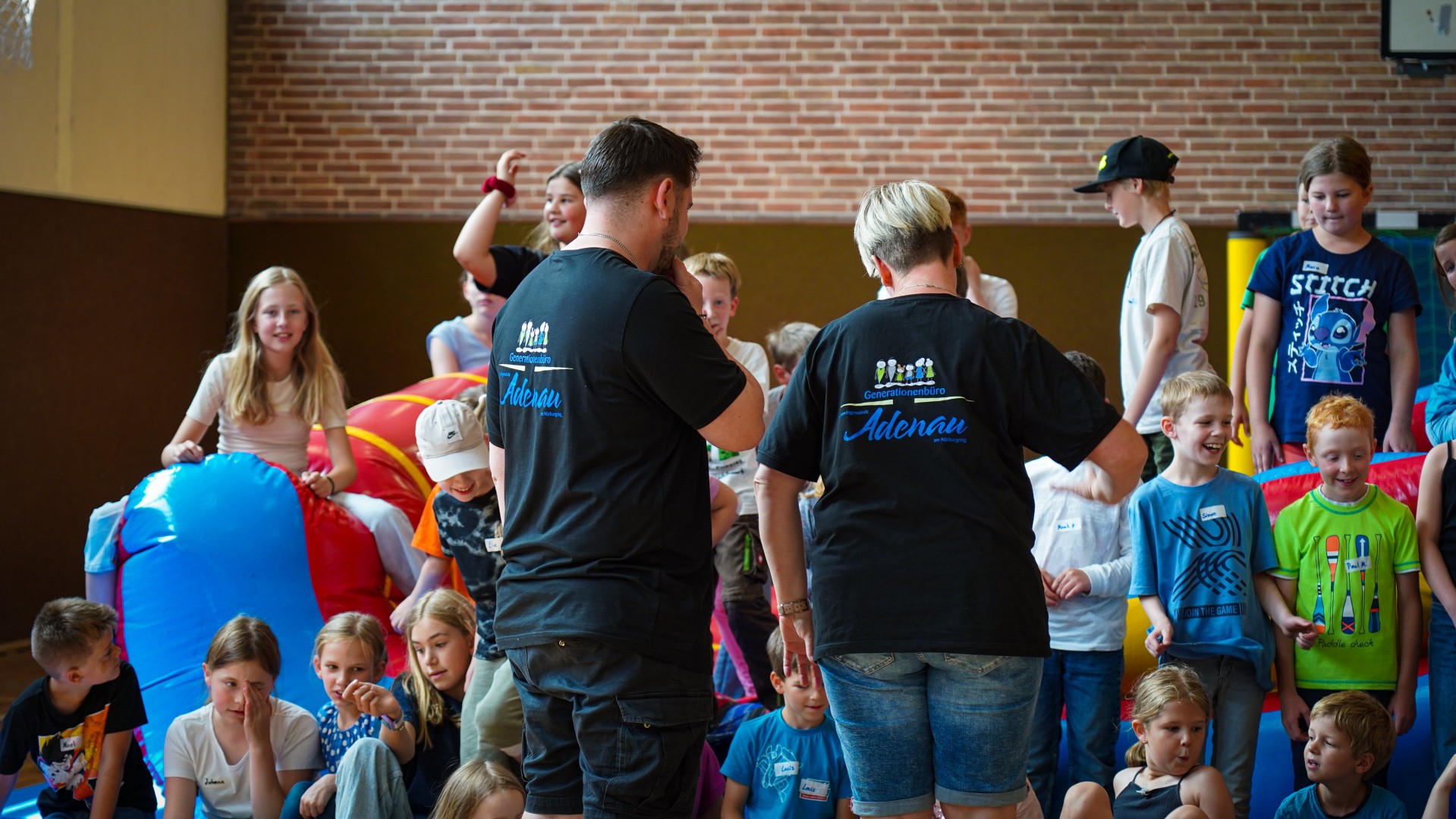In einer Turnhalle sind viele Kinder bei der Ferienfreizeit des Generationenbüros der Verbandsgemeinde Adenau aktiv. Im Vordergrund stehen Jugendpflegerin Silke Schmitz und Jugendpfleger Alexander Schlich mit dem Rücken zur Kamera, beide tragen schwarze T-Shirts mit dem Schriftzug „Generationenbüro Adenau“. Um sie herum spielen und lachen zahlreiche Kinder.