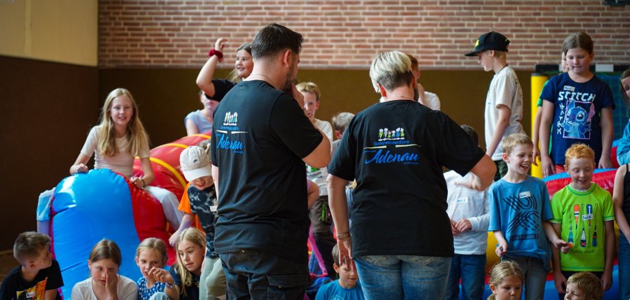 In einer Turnhalle sind viele Kinder bei der Ferienfreizeit des Generationenbüros der Verbandsgemeinde Adenau aktiv. Im Vordergrund stehen Jugendpflegerin Silke Schmitz und Jugendpfleger Alexander Schlich mit dem Rücken zur Kamera, beide tragen schwarze T-Shirts mit dem Schriftzug „Generationenbüro Adenau“. Um sie herum spielen und lachen zahlreiche Kinder.