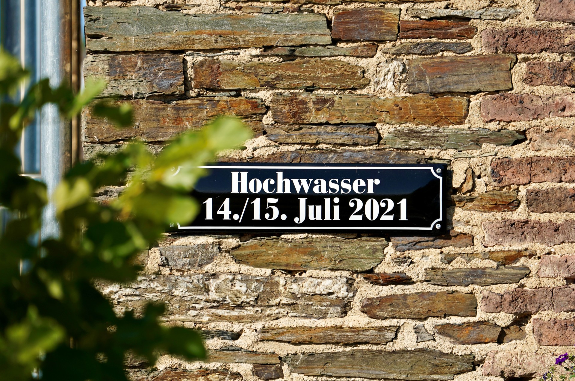 Schwarzes Schild mit weißer Schrift an einer Steinmauer. Text: „Hochwasser 14./15. Juli 2021“.