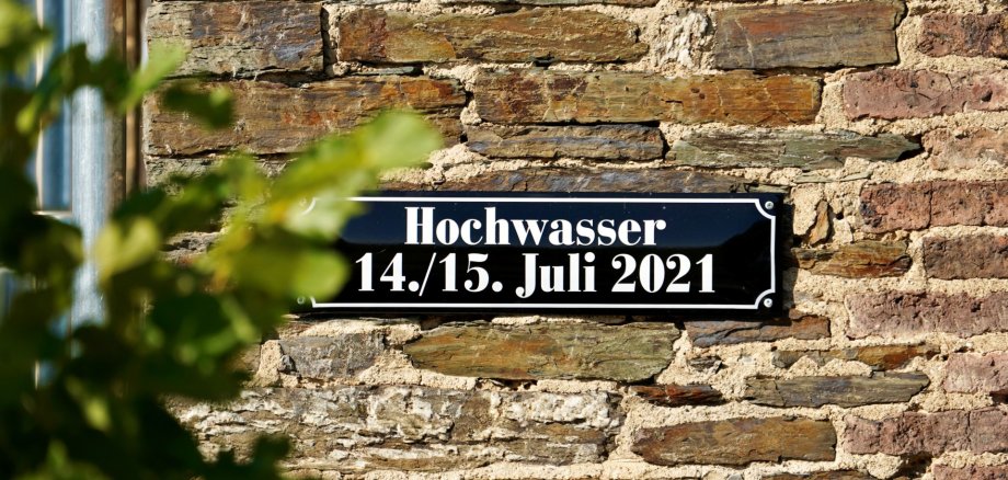 Flutkatastrophe 2021 Schwarzes Schild mit weißer Schrift an einer Steinmauer. Text: „Hochwasser 14./15. Juli 2021“.