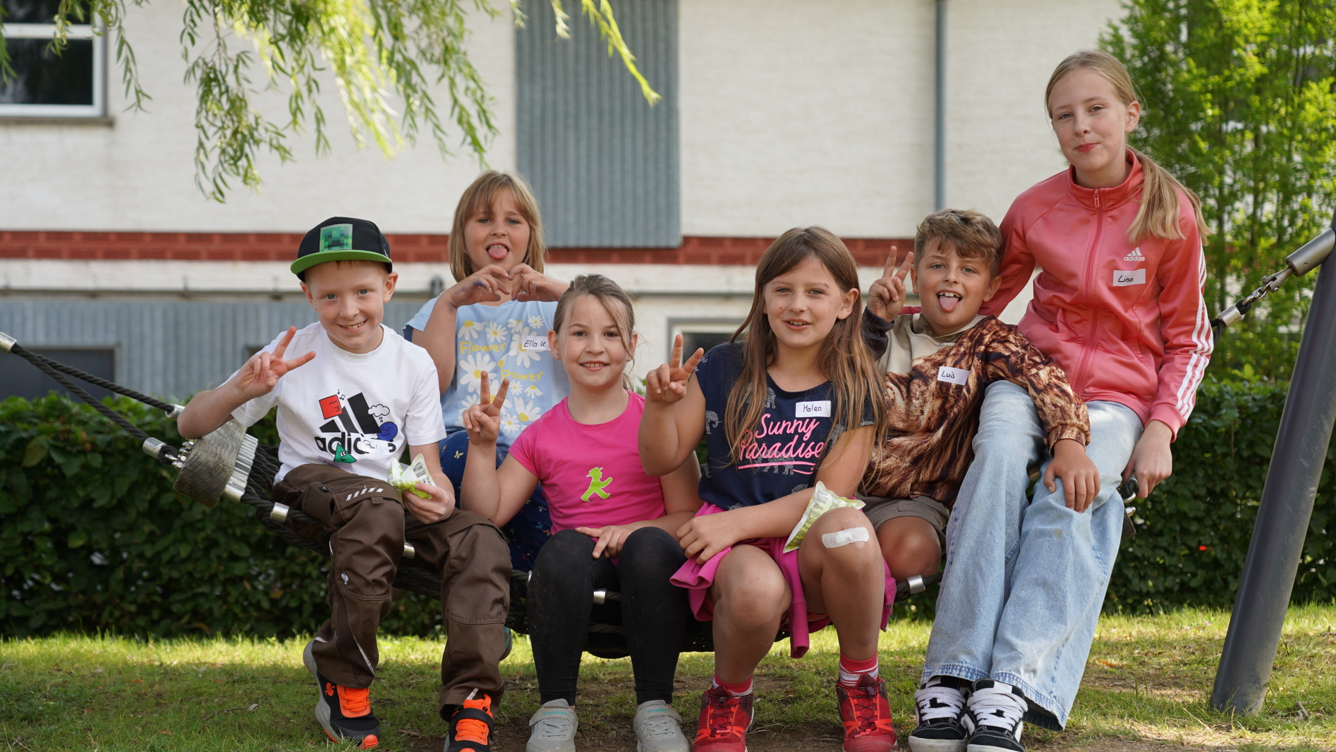 Sechs gut gelaunte Kinder sitzen zusammen in einer Hängematte auf einer Wiese und zeigen „Peace-Zeichen“ und Herzen in die Kamera.