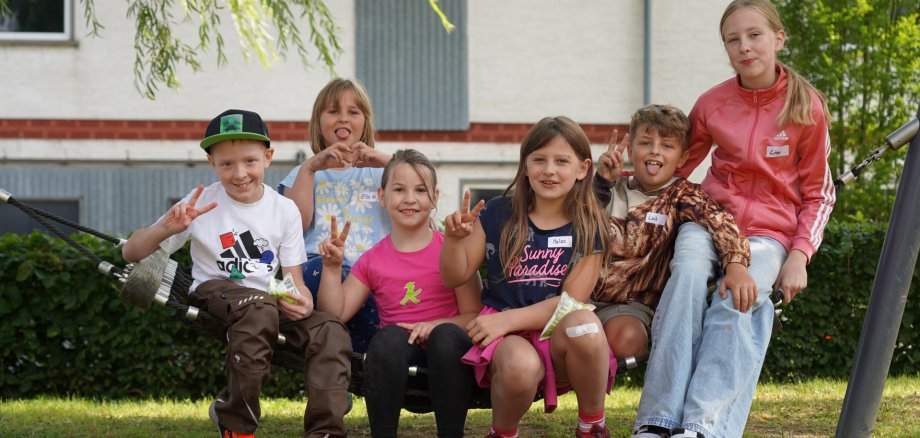 Sechs gut gelaunte Kinder sitzen zusammen in einer Hängematte auf einer Wiese und zeigen „Peace-Zeichen“ und Herzen in die Kamera.