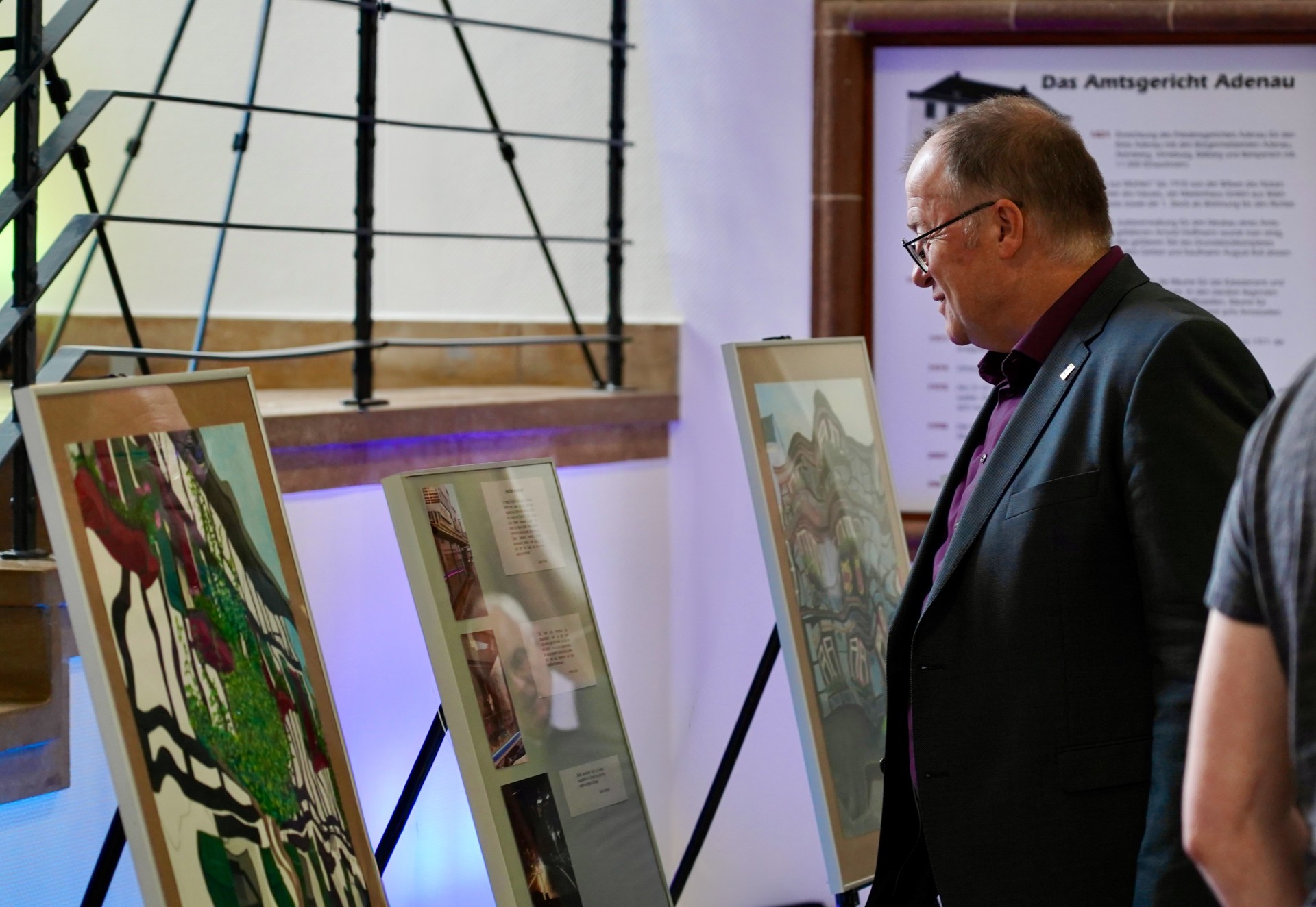 Das Bild zeigt eine Kunstausstellung der Verbandsgemeindeverwaltung Adenau. Im Vordergrund stehen drei Staffeleien mit gerahmten Kunstwerken: links und rechts eine eine verzerrte Darstellung von Gebäuden und in der Mitte eine Collage aus Fotos und Texten. Bürgermeister Guido Nisius betrachtet die Werke aufmerksam.