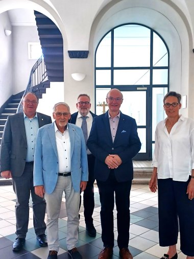 Fünf Personen stehen nebeneinander in einem hellen Raum mit großem Fenster und Treppe im Hintergrund. Von links nach rechts: Bürgermeister der Verbandsgemeinde Adenau, Guido Nisius, die verabschiedete Schiedsperson der Verbandsgemeinde Adenau, Jürgen de Temple, die Schiedsperson der Verbandsgemeinde Mendig, Hans-Jürgen Gross, die ins Amt eingeführte Schiedsperson der Verbandsgemeinde Adenau, Robert Reuter und die Direktorin des Amtsgerichts Bad Neuenahr-Ahrweiler, Dr. Susanne Dreyer-Mälzer.