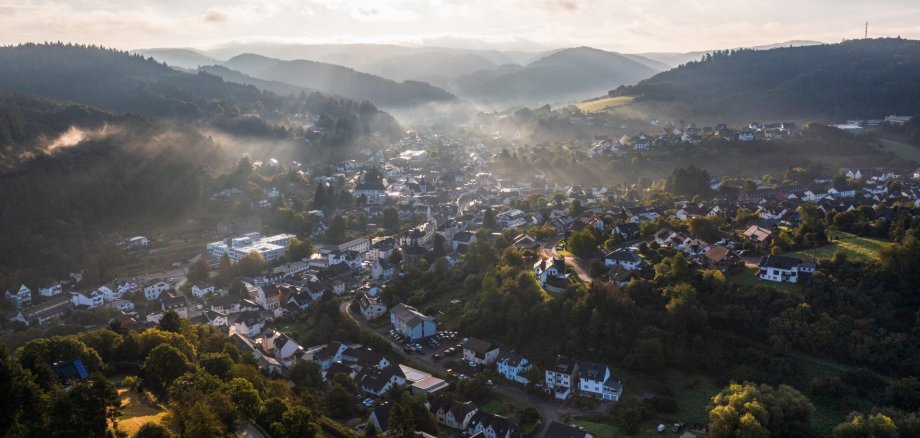 Luftaufnahme der Stadt Adenau, aufgenommen aus Richtung Honerath. Leichte Nebelschwaden ziehen durch das Tal, während die ersten Sonnenstrahlen die Häuser und bewaldeten Hügel rund um die Stadt beleuchten.