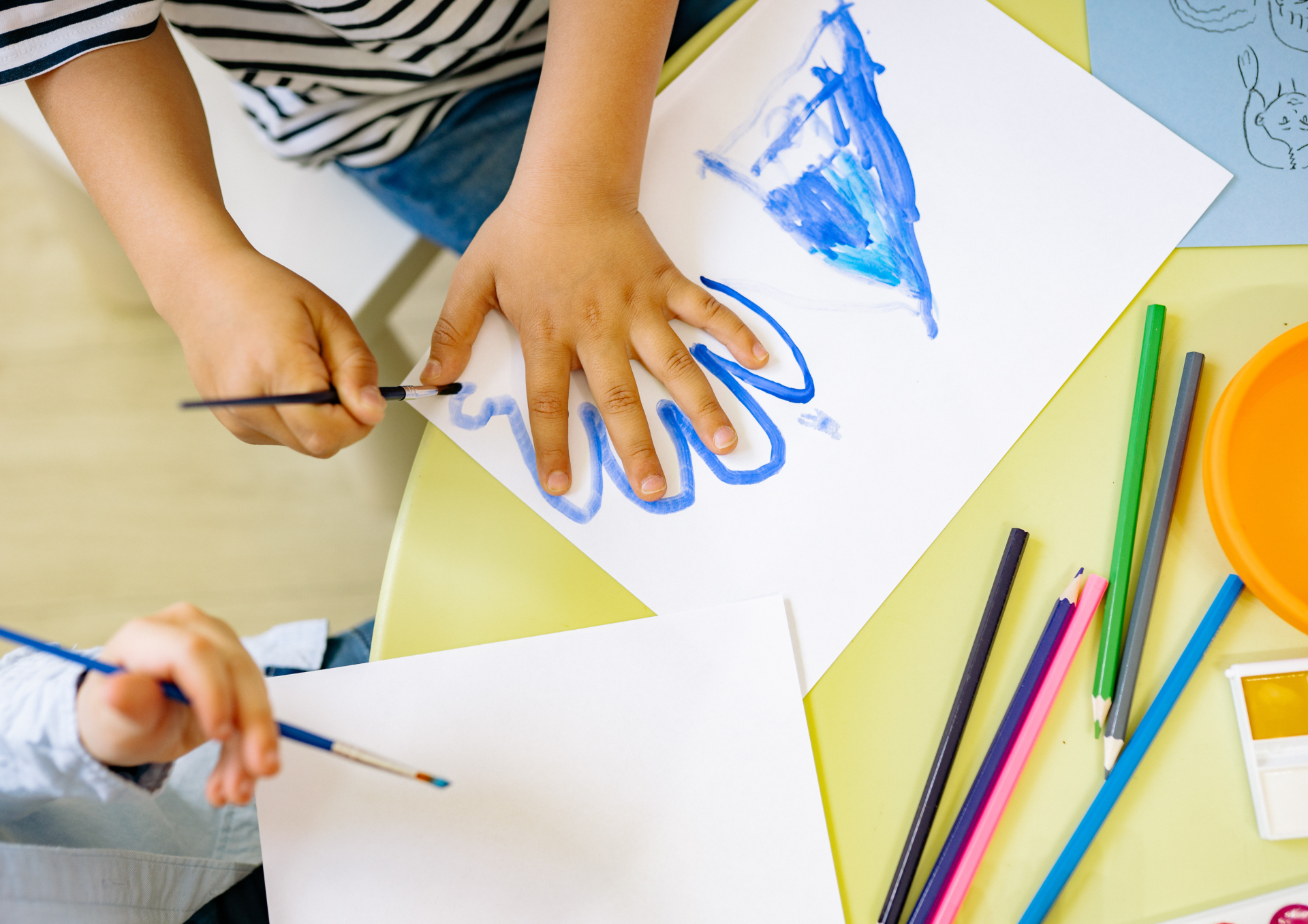 Symbolbild einer Kindertagesstätte: Kinder malen und gestalten gemeinsam Bilder an einem Tisch mit Pinseln, Papier und Buntstiften.