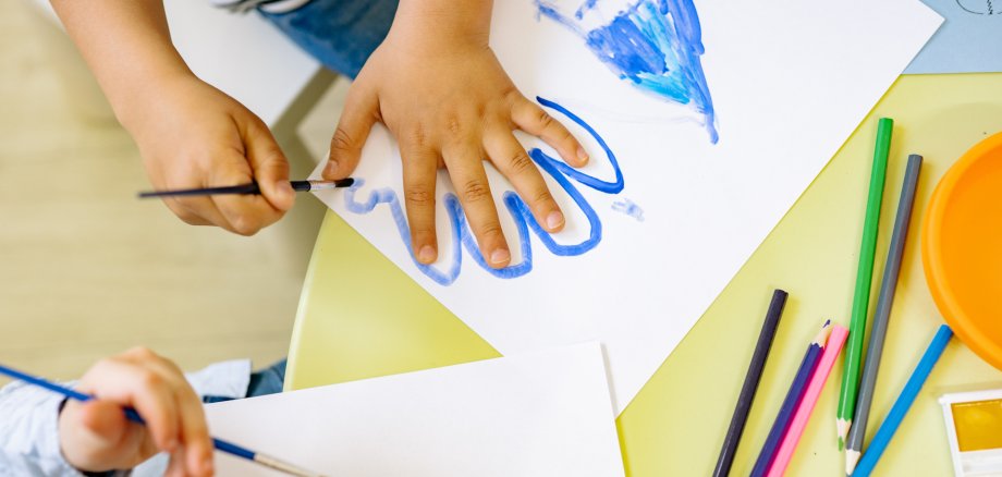 Neubau der Kindertagesstätte Wershofen geht in die Realisierungsphase Symbolbild einer Kindertagesstätte: Kinder malen und gestalten gemeinsam Bilder an einem Tisch mit Pinseln, Papier und Buntstiften.