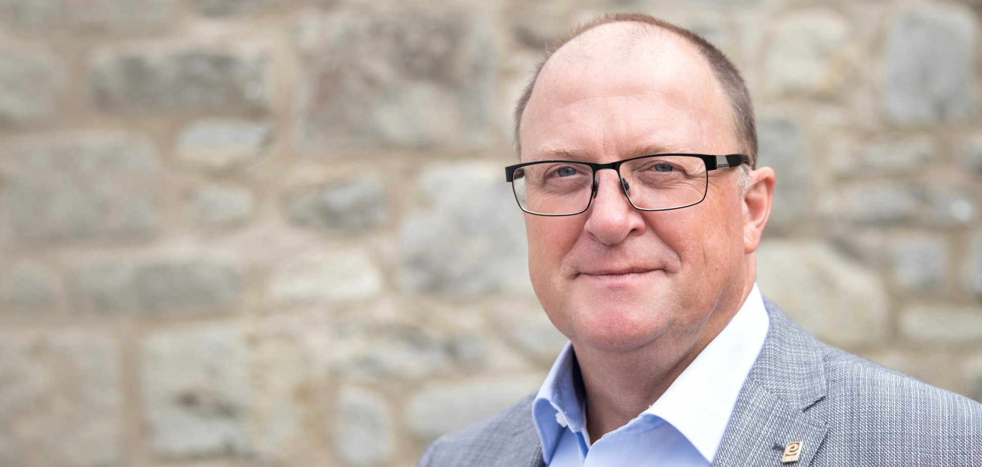 Porträtaufnahme einer Person mit Brille in hellem Sakko und hellblauem Hemd vor einer unscharfen Natursteinmauer. Die Person blickt in die Kamera. Das Foto wird im Zusammenhang mit einer Stellungnahme von Bürgermeister Guido Nisius zu den aktuellen Wald- und Vegetationsbränden verwendet.