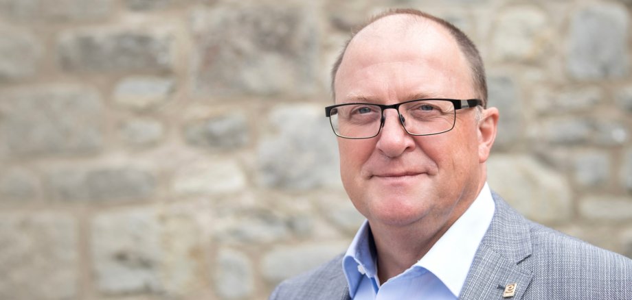 Porträtaufnahme einer Person mit Brille in hellem Sakko und hellblauem Hemd vor einer unscharfen Natursteinmauer. Die Person blickt in die Kamera. Das Foto wird im Zusammenhang mit einer Stellungnahme von Bürgermeister Guido Nisius zu den aktuellen Wald- und Vegetationsbränden verwendet.