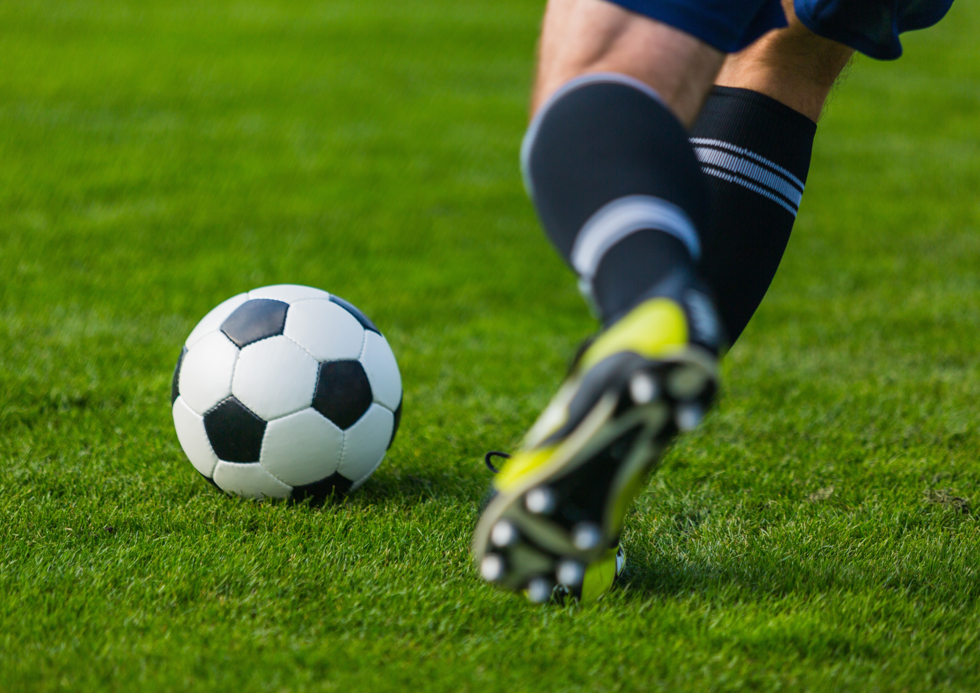 Nahaufnahme eines Fußballs auf einer grünen Rasenfläche. Im Vordergrund nähert sich eine Person in Fußballschuhen dem Ball. Das Bild symbolisiert den Fußballsport und die geplante Modernisierung des Sportplatzes in Wershofen zu einem Naturrasenplatz.