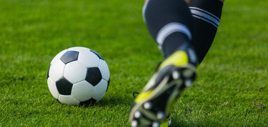 Förderung für die Zukunft des Sports in Wershofen Nahaufnahme eines Fußballs auf einer grünen Rasenfläche. Im Vordergrund nähert sich eine Person in Fußballschuhen dem Ball. Das Bild symbolisiert den Fußballsport und die geplante Modernisierung des Sportplatzes in Wershofen zu einem Naturrasenplatz.