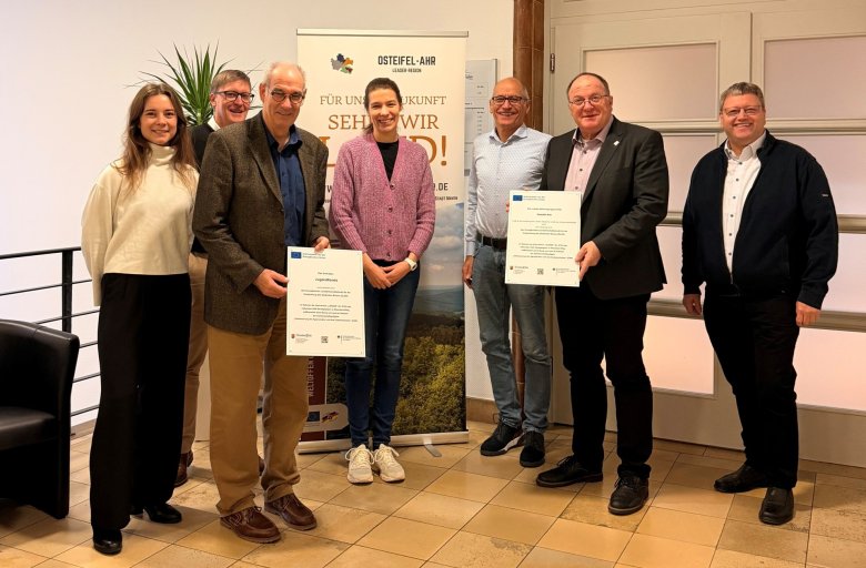 Sieben Personen stehen in einem Innenraum auf einem hellen Fliesenboden und blicken in die Kamera. Zwei Männer halten Zertifikate in den Händen. Hinter der Gruppe hängt ein Banner mit der Aufschrift „OSTEIFEL-AHR – FÜR UNSERE ZUKUNFT SEHEN WIR ROT!“. Die Szene zeigt eine Präsentation der neuen LEADER-Förderhinweis-Schilder durch Vertreter der Verbandsgemeinden und des Regionalmanagements.