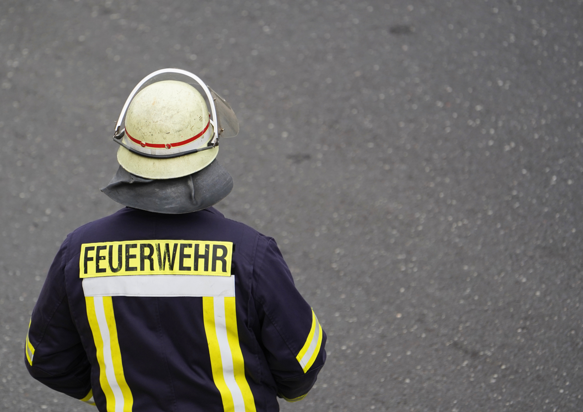Eine Person in dunkelblauer Feuerwehrschutzkleidung steht auf einer grauen Asphaltfläche. Die Person wird von hinten gezeigt und trägt einen hellen Schutzhelm mit Visier sowie eine Schutzhaube. Auf der Jacke befinden sich gelbe Reflexstreifen und der gut sichtbare Schriftzug „FEUERWEHR“. Der Hintergrund besteht ausschließlich aus der Straßenoberfläche.