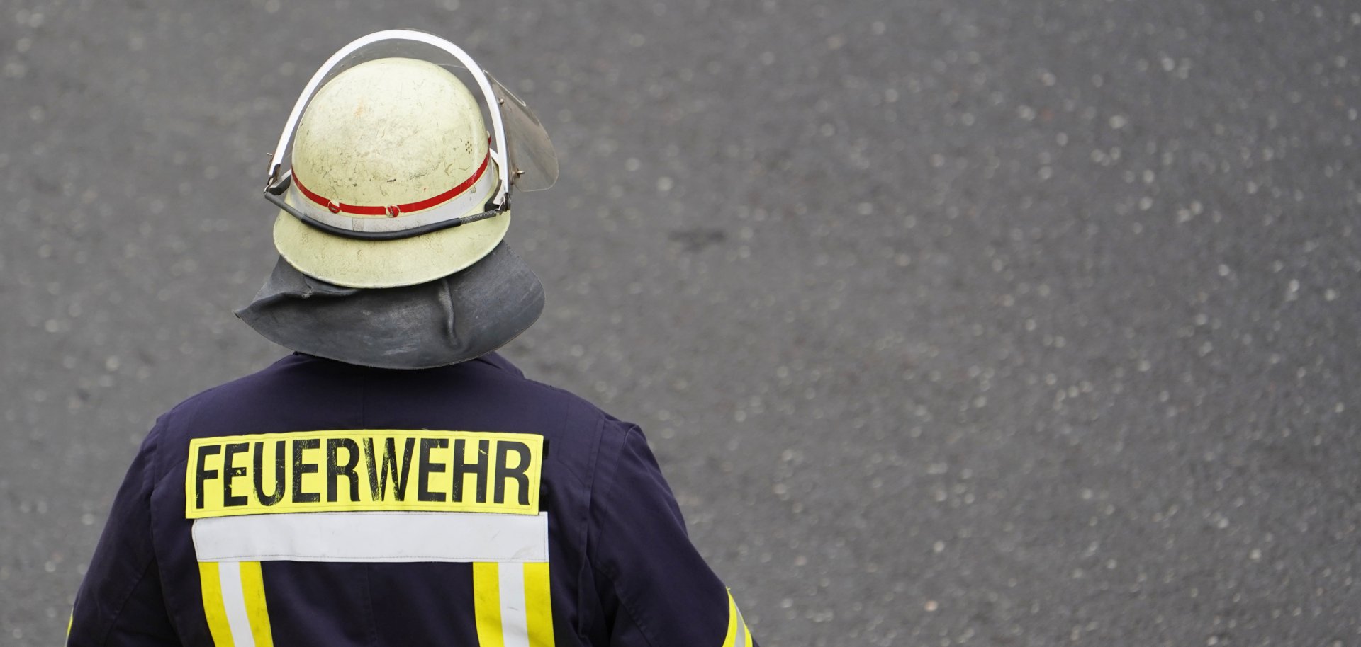 Eine Person in dunkelblauer Feuerwehrschutzkleidung steht auf einer grauen Asphaltfläche. Die Person wird von hinten gezeigt und trägt einen hellen Schutzhelm mit Visier sowie eine Schutzhaube. Auf der Jacke befinden sich gelbe Reflexstreifen und der gut sichtbare Schriftzug „FEUERWEHR“. Der Hintergrund besteht ausschließlich aus der Straßenoberfläche.