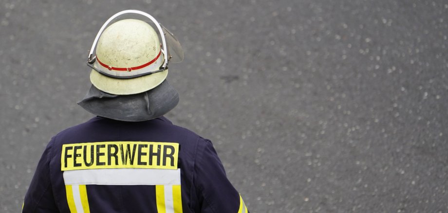 Eine Person in dunkelblauer Feuerwehrschutzkleidung steht auf einer grauen Asphaltfläche. Die Person wird von hinten gezeigt und trägt einen hellen Schutzhelm mit Visier sowie eine Schutzhaube. Auf der Jacke befinden sich gelbe Reflexstreifen und der gut sichtbare Schriftzug „FEUERWEHR“. Der Hintergrund besteht ausschließlich aus der Straßenoberfläche.