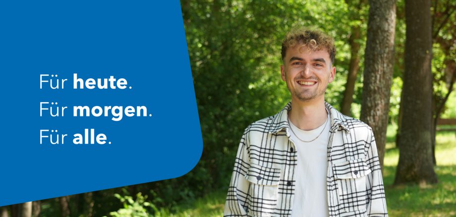 Ein Mann steht lächelnd im Grünen vor Bäumen und Sonnenlicht. Er trägt ein weißes Shirt und eine schwarz‑weiß karierte Jacke. Links im Bild befindet sich ein blauer Bereich mit dem Text „Für heute. Für morgen. Für alle.“.