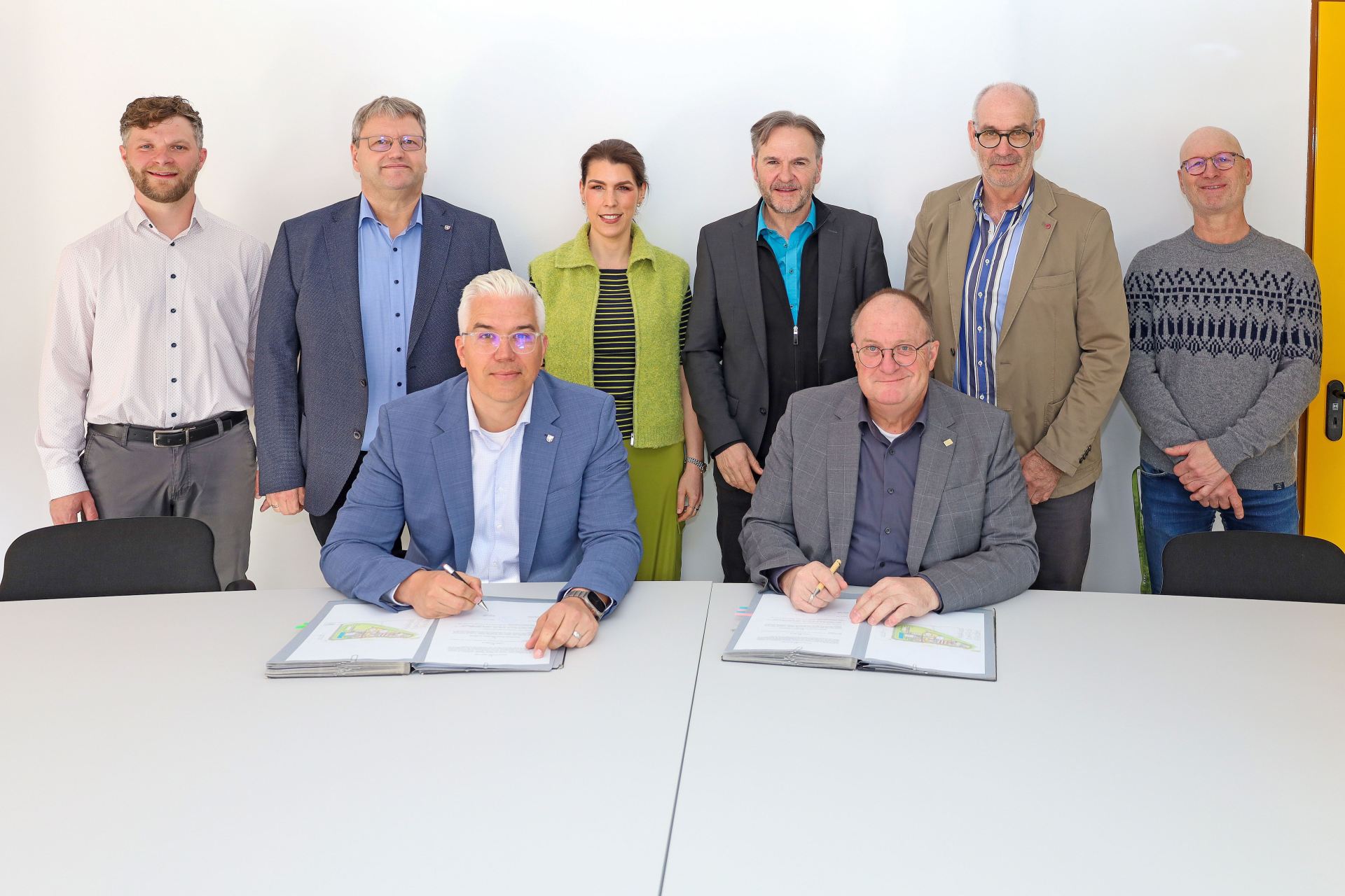 Auf dem Foto sitzen zwei Männer an einem Tisch und unterzeichnen Dokumente. Hinter ihnen stehen sechs weitere Personen in einer Reihe. Die Aufnahme entstand in einem Besprechungsraum der Kläranlage Dümpelfeld und zeigt die Beteiligten der feierlichen Unterzeichnung im Rahmen der Zusammenarbeit der Verbandsgemeinden Adenau und Altenahr.