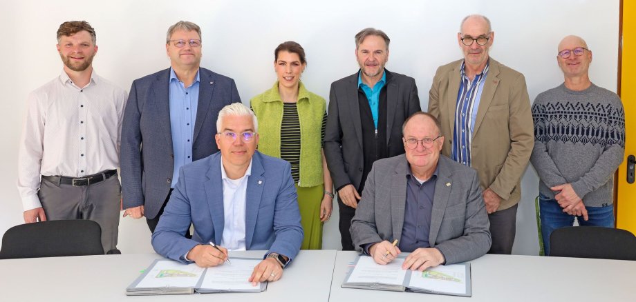 Auf dem Foto sitzen zwei Männer an einem Tisch und unterzeichnen Dokumente. Hinter ihnen stehen sechs weitere Personen in einer Reihe. Die Aufnahme entstand in einem Besprechungsraum der Kläranlage Dümpelfeld und zeigt die Beteiligten der feierlichen Unterzeichnung im Rahmen der Zusammenarbeit der Verbandsgemeinden Adenau und Altenahr.