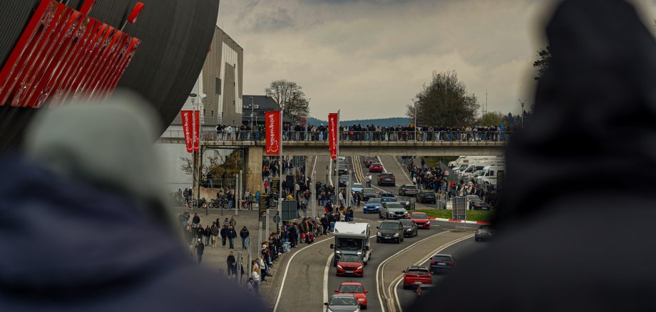 Foto zeigt zwei Personen im Vordergrund von hinten, die auf eine stark befahrene Straße mit vielen Autos und Fußgängern blicken. Im Hintergrund sind zahlreiche Menschen auf einem Fußgängerüberweg und am Straßenrand versammelt. Rote Fahnen mit „Nürburgring“ wehen an Laternenmasten.