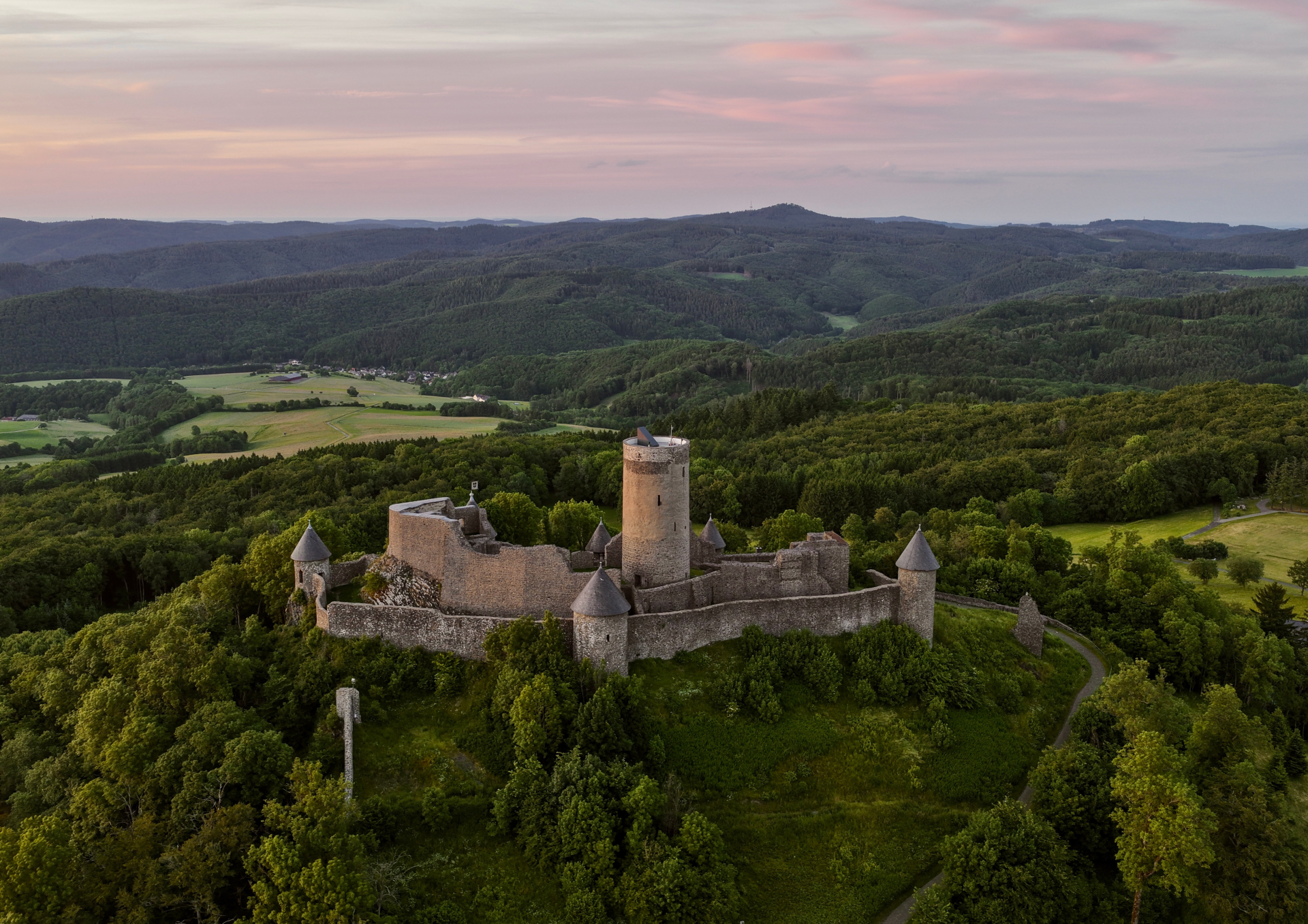 Luftaufnahme der mittelalterlichen Nürburg auf einer Anhöhe in der Eifel. Die Burgruine mit ihrem markanten Rundturm ist von grünen Wäldern und einer weitläufigen Hügellandschaft umgeben. Im Hintergrund erstrecken sich Felder und bewaldete Höhen unter einem Abendhimmel.