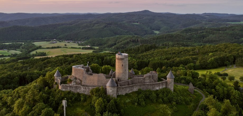 Nürburg lädt in den Sommerferien zum kostenlosen Entdecken ein Luftaufnahme der mittelalterlichen Nürburg auf einer Anhöhe in der Eifel. Die Burgruine mit ihrem markanten Rundturm ist von grünen Wäldern und einer weitläufigen Hügellandschaft umgeben. Im Hintergrund erstrecken sich Felder und bewaldete Höhen unter einem Abendhimmel.
