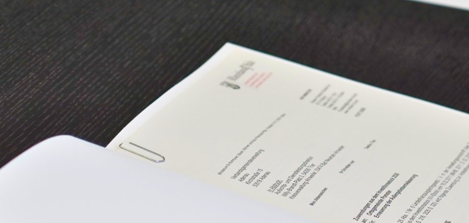 Nahaufnahme eines Förderbescheids des Landes Rheinland-Pfalz auf einem dunklen Tisch. Das Dokument ist mit einer Büroklammer versehen und enthält die Anschrift der Verbandsgemeindeverwaltung Adenau sowie Angaben zur Förderung für die Erneuerung der Außengebietsentwässerung in der Ortsgemeinde Pomster.