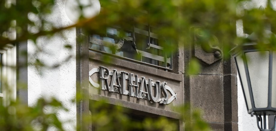 Ansicht eines Rathausgebäudes mit dem Schriftzug „Rathaus“ über einem Eingang. Im Vordergrund sind unscharfe grüne Blätter zu sehen, rechts befindet sich eine Straßenlaterne.