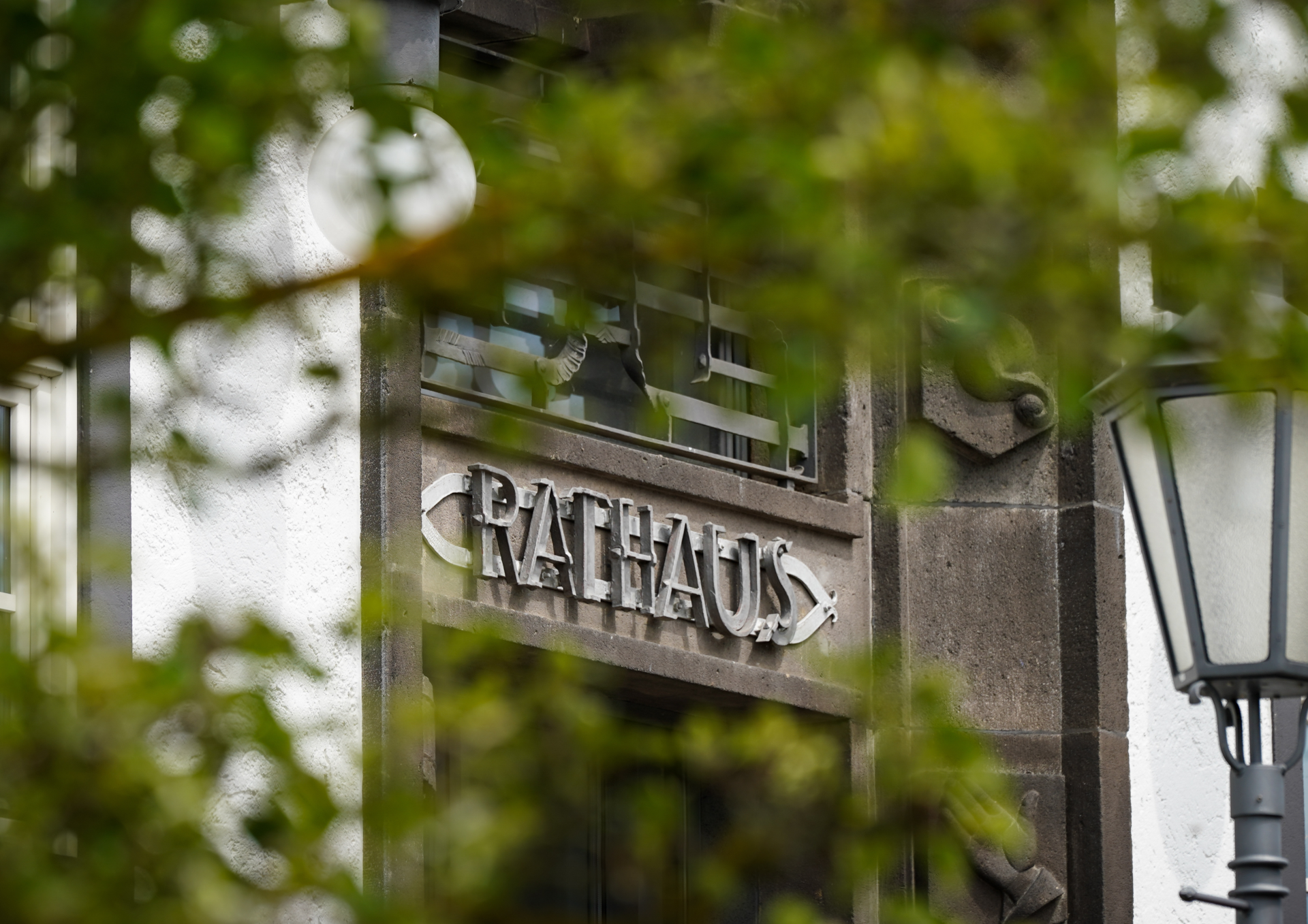 Ansicht eines Rathausgebäudes mit dem Schriftzug „Rathaus“ über einem Eingang. Im Vordergrund sind unscharfe grüne Blätter zu sehen, rechts befindet sich eine Straßenlaterne.