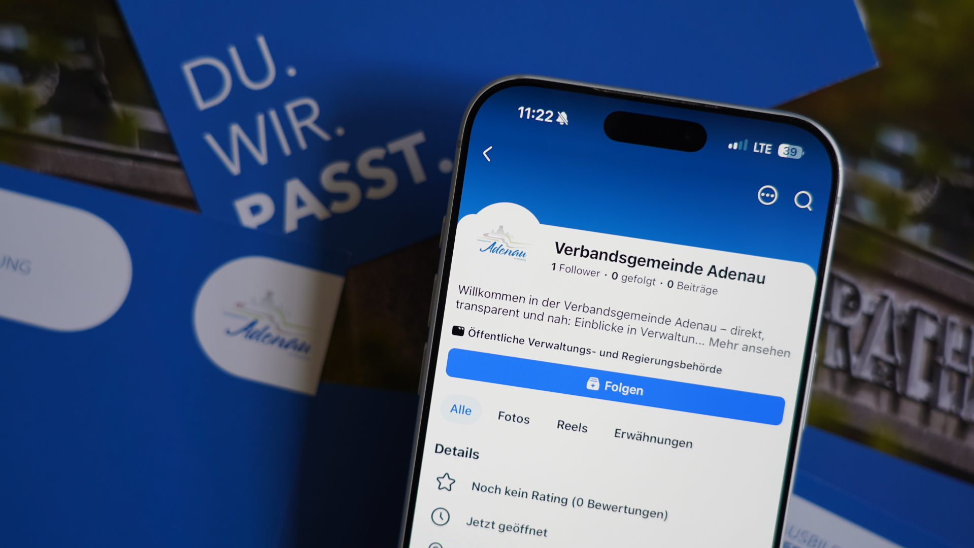 Ein Smartphone zeigt die Facebook-Seite der Verbandsgemeinde Adenau. Hinter dem Smartphone liegen blau‑weiße Broschüren.