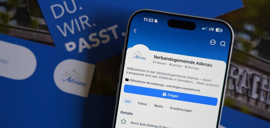 Verbandsgemeinde Adenau erweitert Informationsangebot: Jetzt auch auf Facebook Ein Smartphone zeigt die Facebook-Seite der Verbandsgemeinde Adenau. Hinter dem Smartphone liegen blau‑weiße Broschüren.