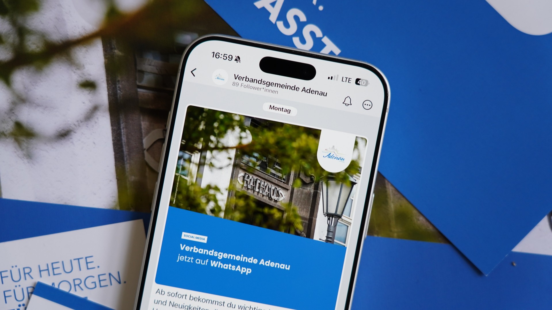 Ein Smartphone zeigt einen Social‑Media‑Post der Verbandsgemeinde Adenau. Der Beitrag informiert darüber, dass die Verwaltung nun auf WhatsApp erreichbar ist. Auf dem Display ist ein Foto des Rathauses mit einem Laternenmast und Baumästen zu sehen. Hinter dem Smartphone liegen blau‑weiße Broschüren.