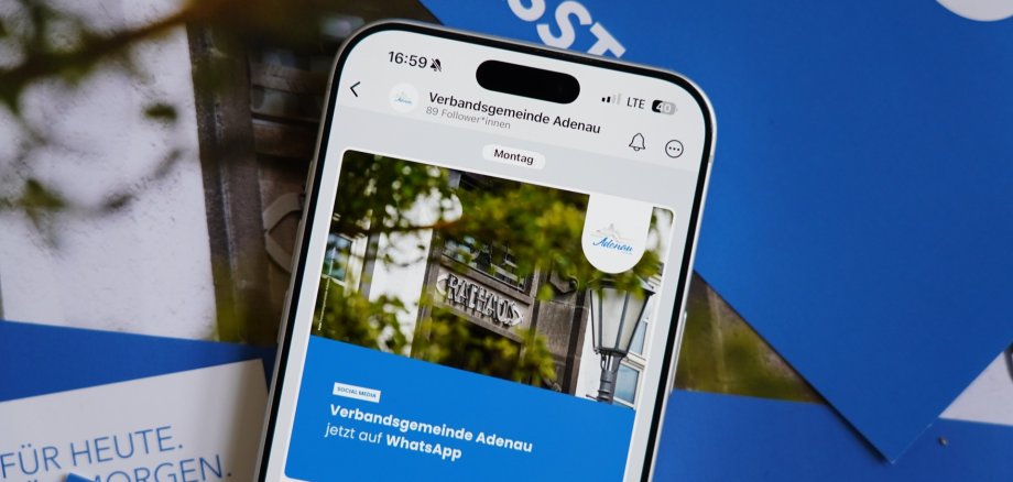 Ein Smartphone zeigt einen Social‑Media‑Post der Verbandsgemeinde Adenau. Der Beitrag informiert darüber, dass die Verwaltung nun auf WhatsApp erreichbar ist. Auf dem Display ist ein Foto des Rathauses mit einem Laternenmast und Baumästen zu sehen. Hinter dem Smartphone liegen blau‑weiße Broschüren.