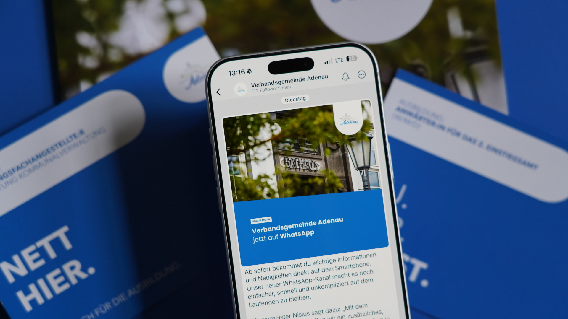 Ein Smartphone zeigt einen Social‑Media‑Post der Verbandsgemeinde Adenau. Der Beitrag informiert darüber, dass die Verwaltung nun auf WhatsApp erreichbar ist. Auf dem Display ist ein Foto des Rathauses mit einem Laternenmast und Baumästen zu sehen. Hinter dem Smartphone liegen blau‑weiße Broschüren.
