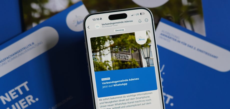 Ein Smartphone zeigt einen Social‑Media‑Post der Verbandsgemeinde Adenau. Der Beitrag informiert darüber, dass die Verwaltung nun auf WhatsApp erreichbar ist. Auf dem Display ist ein Foto des Rathauses mit einem Laternenmast und Baumästen zu sehen. Hinter dem Smartphone liegen blau‑weiße Broschüren.