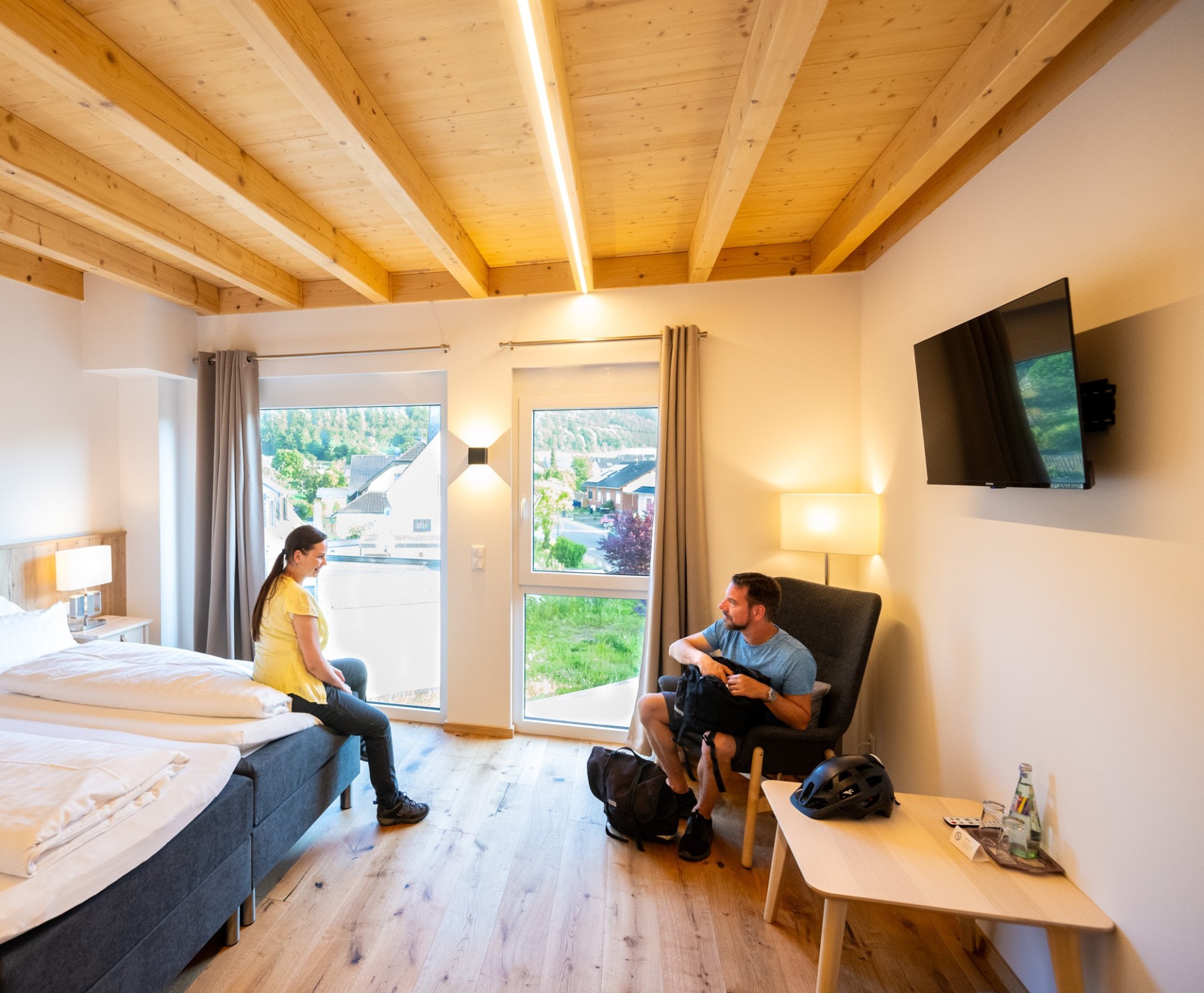 Helles, modern eingerichtetes Gästezimmer mit Holzbalkendecke, zwei Betten, Sesseln und großen Fenstern. Zwei Personen halten sich im Raum auf. Die Aufnahme vermittelt die wohnliche Atmosphäre einer touristischen Unterkunft in der Eifel.
