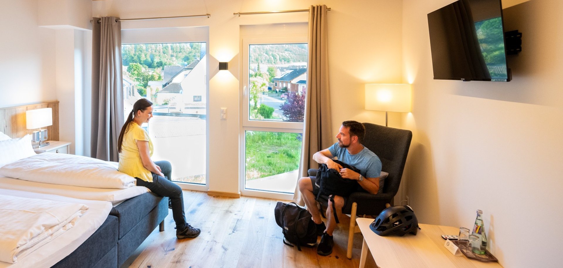 Helles, modern eingerichtetes Gästezimmer mit Holzbalkendecke, zwei Betten, Sesseln und großen Fenstern. Zwei Personen halten sich im Raum auf. Die Aufnahme vermittelt die wohnliche Atmosphäre einer touristischen Unterkunft in der Eifel.