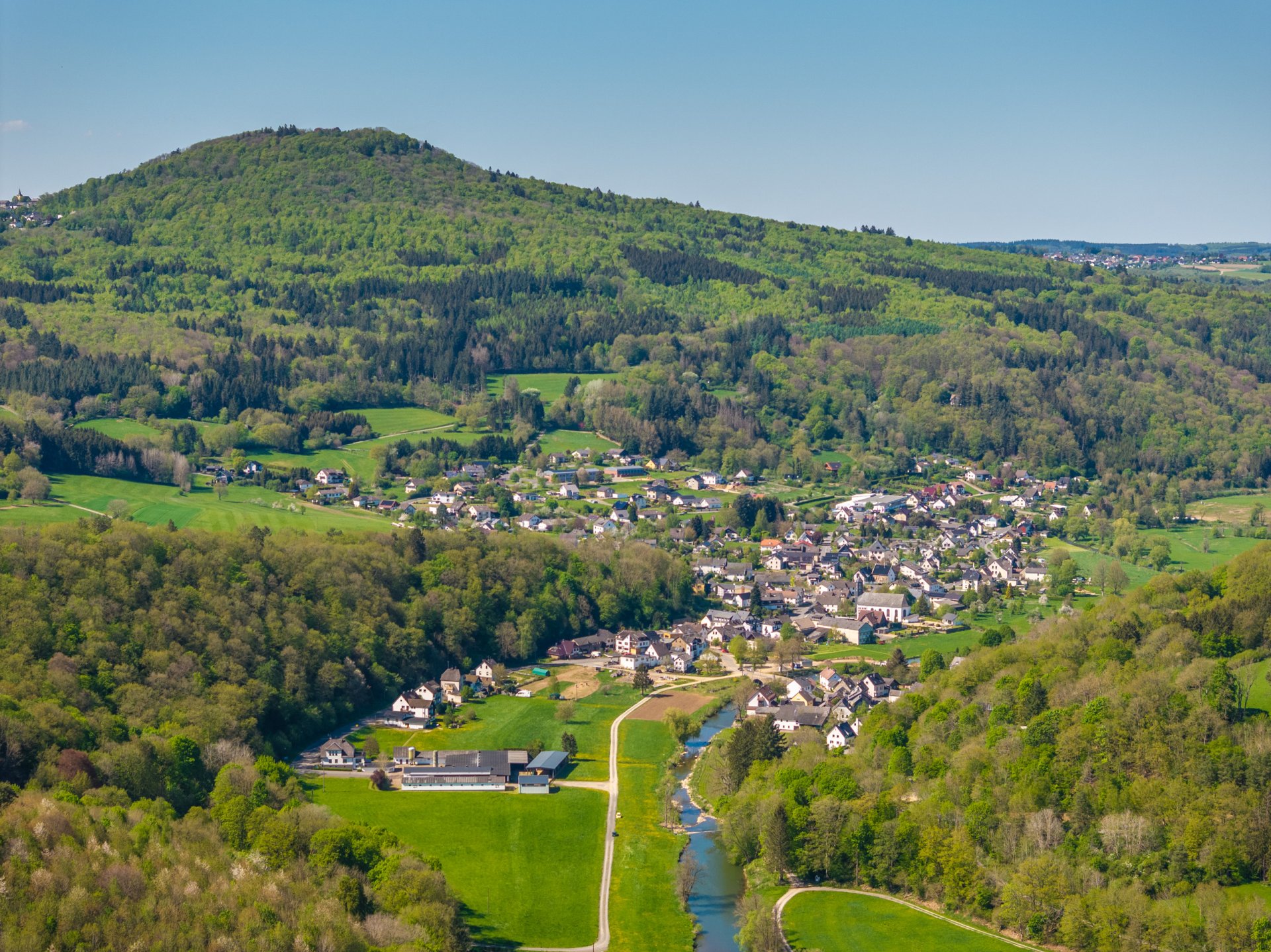 Luftaufnahme des Eifelortes Antweiler, eingebettet in grüne Hügel und Wälder. Durch den Ort fließt ein kleiner Fluss, flankiert von Wiesen und Häusern. Im Hintergrund erhebt sich der bewaldete Aremberg unter klarem blauem Himmel.
