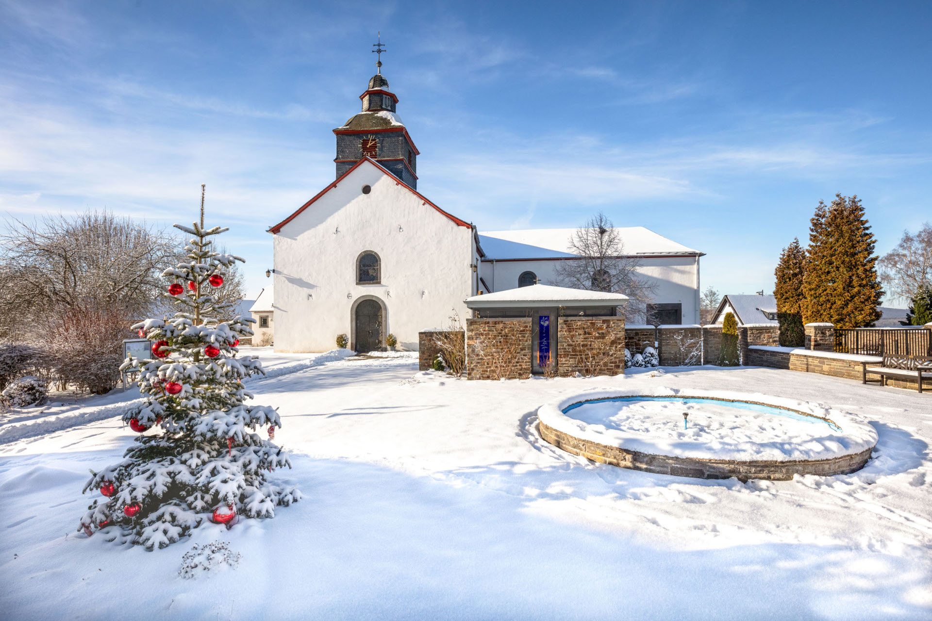Schneebedeckte Kirche in Barweiler mit geschmücktem Weihnachtsbaum und rundem Brunnen unter blauem Himmel