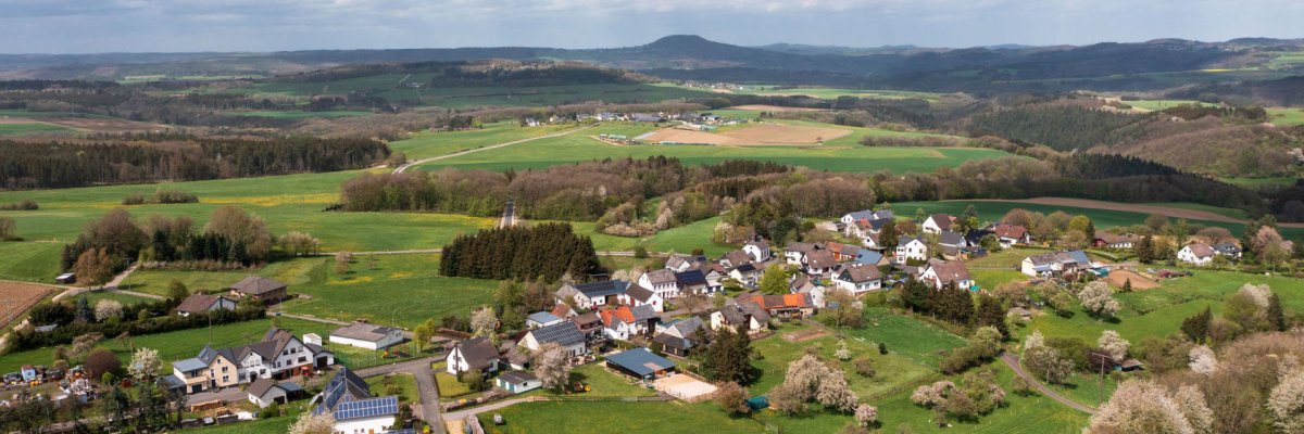 Luftaufnahme des Orts Dankerath mit Häusern in verschiedenen Dachfarben, eingebettet in grüne Felder und sanfte Hügel. Straßen verbinden die Gebäude, und die Landschaft erstreckt sich unter einem teils bewölkten Himmel mit Wäldern und Wiesen bis zum Horizont.
