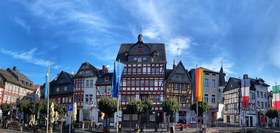 Straßenszene mit traditionellen Fachwerkhäusern in Weiß, Rot und Braun. Mehrere Nationalflaggen – darunter Deutschland, Frankreich, Italien und eine blaue Flagge – hängen an Masten entlang der Straße. Bäume und Außensitzbereiche von Cafés säumen den Gehweg. Himmel mit Schleierwolken.