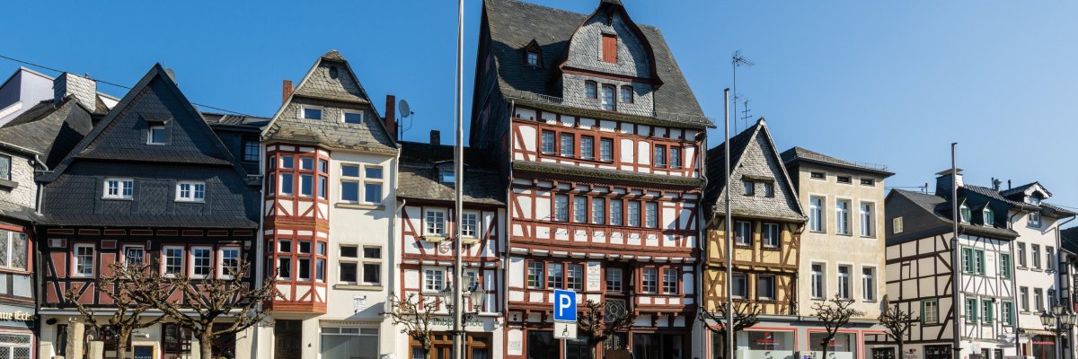 Reihe traditioneller Fachwerkhäuser mit steilen Dächern, vielen Fenstern und dekorativen Holzelementen. Fußgängerbereich mit Bänken und geformten Bäumen, Parkplatzschild im Vordergrund. Klarer blauer Himmel
