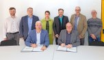 Auf dem Foto sitzen zwei Männer an einem Tisch und unterzeichnen Dokumente. Hinter ihnen stehen sechs weitere Personen in einer Reihe. Die Aufnahme entstand in einem Besprechungsraum der Kläranlage Dümpelfeld und zeigt die Beteiligten der feierlichen Unterzeichnung im Rahmen der Zusammenarbeit der Verbandsgemeinden Adenau und Altenahr.